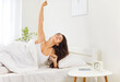 © Studio Romantic - Cheerful young brunette woman waking up early in morning in her bedroom with alarm clock and smartphone on table. Happy smiling Caucasian girl stretching in her comfortable bed after good night sleep.