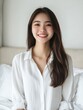 © The 2R Artificiality - Joyful Asian woman with long dark hair smiling in a bright white bedroom, exuding happiness and success with ample empty space for text.
