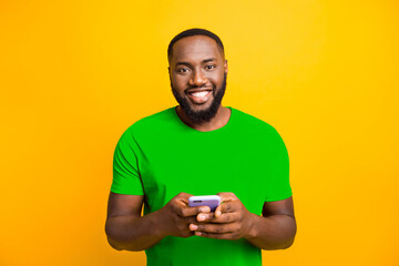 Wall Mural - Photo of simple good cute handsome black man holding his phone with both hands smiling at camera while isolated with yellow background