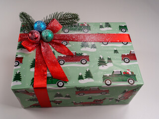 A beautifully wrapped gift features colorful paper with vintage truck designs and winter scenes. Adorned with a sparkling red ribbon and ornaments, it celebrates the holiday spirit.