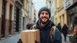 © Ivan - A young man with a beard and beanie joyfully holds a cardboard package as he walks down a narrow city street. Colorful buildings and pedestrians surround him during the daytime