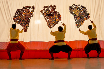 Wall Mural - Three player men wear traditional cloth with golden shade stand and perform grand shadow play in front of white screen with reflection on the floor.