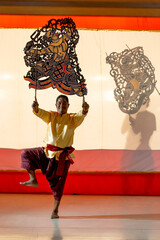 Wall Mural - Vertical image of Asian senior man stand with one leg to perform grand shadow play in front of his team stay behide in the shadow of other charactor craft.