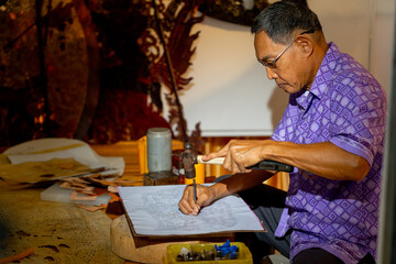 Wall Mural - Side view of Asian senior man wear glasses use hammer and chisel to create grand shadow play craft and he concentrate to work in area of workplace