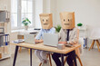 © Studio Romantic - Busy office workers sitting at table, wearing paper bag with drawn face looking at laptop screen, reading message email with news, chatting with clients online, working remotely, business routine