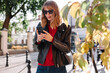 © Viktor Solomin/Stocksy - Smiling woman in sunglasses checking her phone in autumn on street