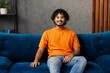 © ViDi Studio - Young smiling happy cheerful Indian man wear orange casual clothes sits on blue sofa couch look camera stay at home hotel flat rest relax spend free spare time in living room indoor. Lounge concept.