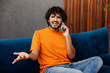 © ViDi Studio - Young sad Indian man wear orange casual clothes sits on blue sofa couch talk speak on mobile cell phone stay at home hotel flat rest relax spend free spare time in living room indoor. Lounge concept.