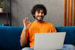 © ViDi Studio - Young IT Indian man wear casual clothes sit on blue sofa couch hold use work on laptop pc computer show ok okay stay at home hotel flat rest relax spend free time in living room indoor Lounge concept