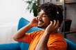 © ViDi Studio - Close up young Indian man wear orange casual clothes sits on blue sofa couch listen music in headphones stay at home hotel flat rest relax spend free spare time in living room indoor. Lounge concept.