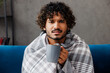 © ViDi Studio - Young sick ill exhausted tired Indian man he wrapped in grey plaid sits on blue sofa couch drink hot tea stay at home hotel flat rest relax spend free spare time in living room indoor. Lounge concept.