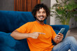 © ViDi Studio - Young Indian man wear orange casual clothes sit near blue sofa couch hold use point on mobile cell phone stay at home hotel flat rest relax spend free spare time in living room indoor. Lounge concept.