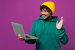 © ViDi Studio - Young happy Indian IT man wear green hoody yellow hat casual clothes hold use work on laptop pc computer waving hand talk speak isolated on plain purple background studio portrait. Lifestyle concept.