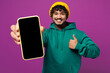 © ViDi Studio - Young Indian man he wear green hoody yellow hat casual clothes hold in hand use mobile cell phone with blank screen workspace area show thumb up isolated on plain purple background. Lifestyle concept.