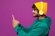 © ViDi Studio - Side view young happy Indian man wear green hoody yellow hat casual clothes listen to music in headphones use mobile cell phone isolated on plain purple background studio portrait. Lifestyle concept.