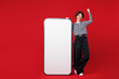 © ViDi Studio - Full body young cool woman in blue striped sweatshirt casual clothes big huge blank screen mobile cell phone smartphone with copy space mockup area do winner gesture isolated on plain red background