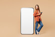 © ViDi Studio - Full body young woman she wear orange shirt casual clothes big huge blank screen mobile cell phone with workspace copy space mockup area use smartphone look aside isolated on plain beige background.