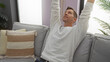 © Krakenimages.com - Young man relaxing indoors with arms raised on a comfortable sofa in a cozy living room apartment filled with soft pillows and natural light.