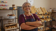 © Krakenimages.com - Woman standing in a bakery with arms crossed, smiling confidently, surrounded by shelves of fresh bread and pastries, wearing glasses and a purple shirt with an apron, clock in the background
