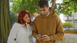 © Krakenimages.com - Man showing smartphone to woman in a park setting surrounded by urban greenery, both focused and engaged, showcasing a moment of modern technology and communication outdoors.