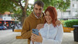 © Krakenimages.com - Woman and man smiling at phone in city park with trees around them, capturing a joyful moment together outdoors, showcasing love and happiness.