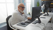 © Krakenimages.com - Man with grey hair and beard in office holding shoulder in pain while working on a computer.
