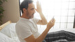 © Krakenimages.com - Middle-aged hispanic man sits on bed in bedroom looking surprised at his mobile phone, holding one hand up in shock.