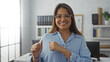 © Krakenimages.com - Hispanic woman smiling in an office while pointing her thumbs backward, showcasing a positive and inviting corporate environment