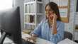 © Krakenimages.com - Smiling young woman talking on the phone while working on a computer in a modern office environment, featuring a neat workstation with bookshelves and a bulletin board.