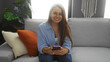 © Krakenimages.com - Young woman sitting on a couch in a living room, smiling and using a smartphone, with a cozy home background.