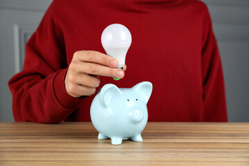 Wall Mural - Man holding light bulb above piggy bank at wooden table, closeup. Energy saving concept