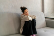 © Mihail - A woman with a bun is sitting on a grey sofa, holding her face in her hand, expressing sadness and distress in a calm, minimalistic room
