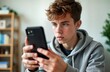© Pete - Teenager using smartphone indoors. Young person looks intently at mobile phone. Casual attire. Modern device in home setting. Possible activities include gaming, messaging, social media. Focus on