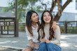 © crizzystudio - Two young female asian students are sitting together on campus, holding notebooks and laughing, enjoying their friendship and academic life