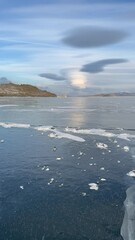 Wall Mural - Vertical video of Baikal Lake in December. Unusual lenticular (Lat. Lenticularis) or lens-shaped clouds over clear ice surface with cracks of frozen Kurkut Bay of Small Sea. Scenic winter landscape