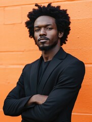 Wall Mural - Stylish African American man in a black fashion jacket posing confidently with crossed arms against a vibrant orange wall showcasing empty copyspace for text