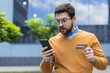 © Tetiana - A young serious man is standing on the street, holding a credit card, looking at the mobile phone in surprise and concern