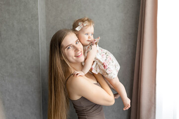 Smiling mother holds her baby daughter close, both dressed in casual attire, warmth and connection in serene home environment. natural lighting enhances tender family moment.