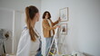 © stockbusters - Man woman decorating wall placing artwork in living room. Husband on ladder