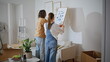 © stockbusters - Spouses decorating living room by arranging paintings on white wall. Happy woman
