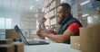 © Framestock - Portrait of African American post office employee scanning parcel with barcode scanner, using laptop, looking at camera. Sorting center workers checking boxes for delivery. Warehouse of online store.