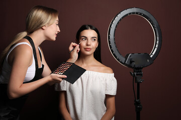 Wall Mural - Professional makeup artist working with beautiful young woman against brown background. Using ring lamp