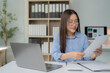 © Tj - Asian businesswoman wearing glasses and a blue shirt, working diligently at her office desk, reviewing documents and analyzing data on her laptop, showcasing a professional environment