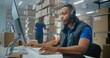 © Framestock - African American logistics coordinator wearing headset chats with customer on call, uses computer, works in post office or E-commerce store storage. Sorting center workers carrying cardboard boxes.