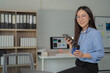 © Tj - Young asian businesswoman wearing glasses, holding smartphone and drinking coffee while sitting on desk in modern office, smiling and looking at camera