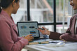 © amnaj - Two businesswomen are discussing a summary report displayed on a tablet screen during a meeting in a modern office