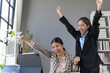 © amnaj - Two euphoric businesswomen celebrating a successful project, raising their arms in victory in a modern office