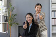© amnaj - Two Asian businesswomen collaborating in a modern office, smiling and giving thumbs up, embodying teamwork and professional success