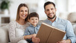 © mirifadapt - smiling couple of parents sitting on sofa with child boy, man holding big book in hands