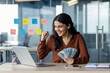 © Liubomir - Young businesswoman investor inside office holding money cash dollars in hands, woman celebrating victory and success looking at laptop screen, received income big win.
